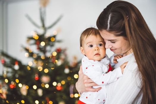 Portrait of young mom and her daughter, baby girl having fun on Christmas, decorating the Christmas tree in festive outfits. Smiling baby girl playing with her mom. 