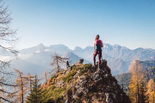 Scenic view of autumn mountain, European Alps, from a view point, where caucasian woman hiker is standing. Sun is shining high up in the mountains, a light mist in the valleys down bellow. Woman mountaineer enjoying the view of majestic Alps on a sunny autumn day. 