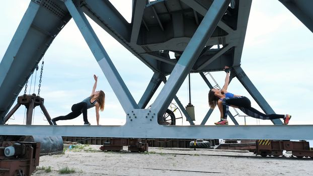 Two young, athletic women, perform strength exercises.They stand on the metal beam of a freight crane. fitness at dawn, on the beach in a cargo port. High quality photo