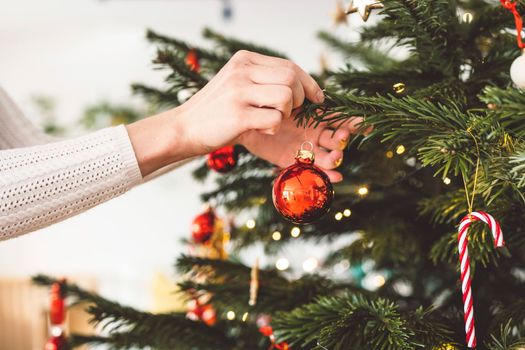 Unrecognizable caucasian woman decorating the Christmas tree, putting red ornament on the tree, holding a red Christmas ornament. 