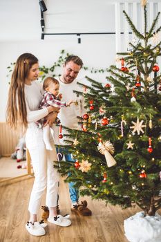 Happy young cheerful caucasian family of three mom dad and baby girl having fun decorating the Christmas tree. Family Christmas portrait. 