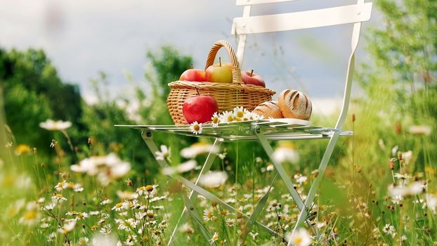In the middle of a chamomile lawn, near a forest, against a blue sky, stands a white chair, on it a composition of a basket with red apples and bread, and a bouquet of daisies stands. High quality photo