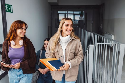 Group of three friends, young caucasian women students walking inside the school during a break, headed to another classroom. Students carrying their books with them. 