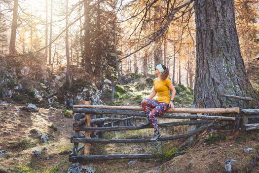 Young caucasian woman hiker sitting on a wooden fence somewhere in the autumn forest while on a hike, wearing a bright yellow shirt and colorful leggings. 