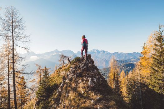 Scenic view of autumn mountain, European Alps, from a view point, where caucasian woman hiker is standing. Sun is shining high up in the mountains, a light mist in the valleys down bellow. Woman mountaineer enjoying the view of majestic Alps on a sunny autumn day. 