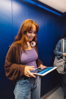 Vertical photo of young caucasian woman with handphones around her neck, holding a dgital tablet while standing in an elevator.