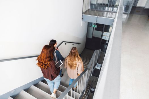 Group of three friends, young caucasian women students walking inside the school during a break, headed to another classroom. Students carrying their books with them. 