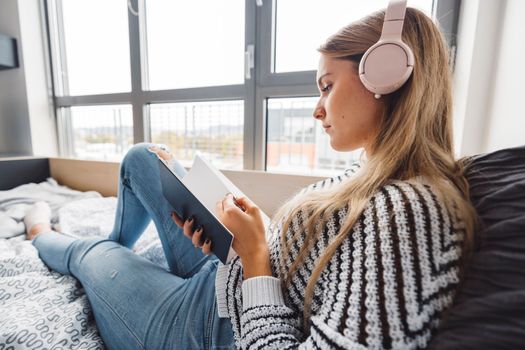 Blonde young caucasian woman lying on her dorm room bed next to a window putting on headphones, listening to music on a cold cloudy autumn day.