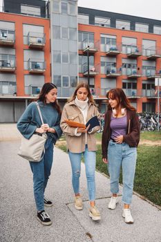 Group of three cheerful female collage students outside their dorm on a cold autumn day, walking on campus to get to their class. 