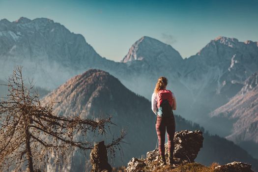 Scenic view of autumn mountain, European Alps, from a view point, where caucasian woman hiker is standing. Sun is shining high up in the mountains, a light mist in the valleys down bellow. Woman mountaineer enjoying the view of majestic Alps on a sunny autumn day. 