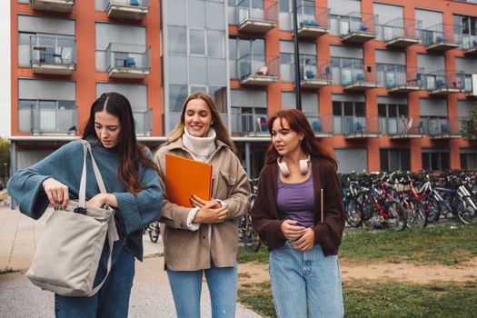 Group of three cheerful female collage students outside their dorm on a cold autumn day, walking on campus to get to their class. 