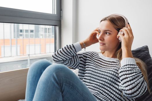 Blonde young caucasian woman lying on her dorm room bed next to a window putting on headphones, listening to music on a cold cloudy autumn day.