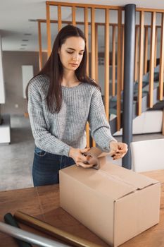 Young caucasian woman wrapping a gift, getting it ready to send in the post. Woman packaging a surprise gift for a family member, packaging it into a brown cardboard box.