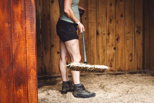 Unrecognizable woman cleaning horses stables in working ger on a hot summer day. 