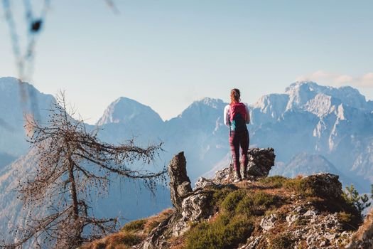 Scenic view of autumn mountain, European Alps, from a view point, where caucasian woman hiker is standing. Sun is shining high up in the mountains, a light mist in the valleys down bellow. Woman mountaineer enjoying the view of majestic Alps on a sunny autumn day. 