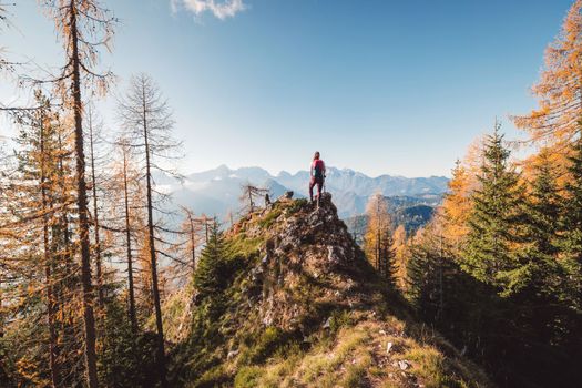 Scenic view of autumn mountain, European Alps, from a view point, where caucasian woman hiker is standing. Sun is shining high up in the mountains, a light mist in the valleys down bellow. Woman mountaineer enjoying the view of majestic Alps on a sunny autumn day. 