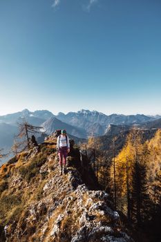 Scenic view of autumn mountain, European Alps, from a view point, where caucasian woman hiker is standing. Sun is shining high up in the mountains, a light mist in the valleys down bellow. Woman mountaineer enjoying the view of majestic Alps on a sunny autumn day. 