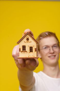 Affordable Housing for Youth Concept. A positive Young European Blond Man holds in his hand a wooden model of house. High quality photo.