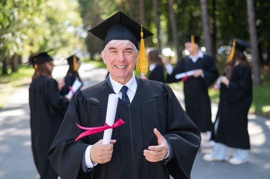 A group of graduates in robes outdoors. An elderly student rejoices at receiving a diploma