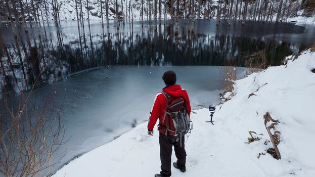 A guy standing on the shore admires a mountain lake. From here you can see the mirror-black color of the water, which reflects snowy mountains, green forest, clouds. Kaindy Lake is freezing Kazakhstan