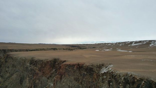 A large crack in the ground overlooking the snowy hills. Grey sky covered with clouds. The Grand Canyon in the middle of the steppe. The river runs. Top view from a drone. Kazakhstan, Black Canyon