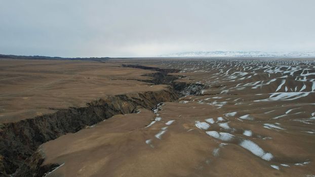 A large crack in the ground overlooking the snowy hills. Grey sky covered with clouds. The Grand Canyon in the middle of the steppe. The river runs. Top view from a drone. Kazakhstan, Black Canyon