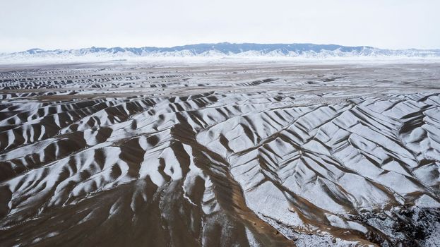 The high hills are like waves covered with snow. Black mountains with white snow are visible in the distance. Grey sky with clouds. Top view from the drone on patterns of the earth. Dark brown stones