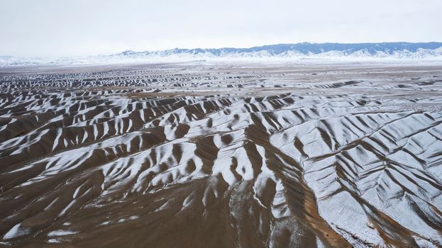 The high hills are like waves covered with snow. Black mountains with white snow are visible in the distance. Grey sky with clouds. Top view from the drone on patterns of the earth. Dark brown stones