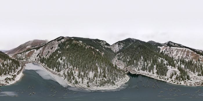 Kaindy Mountain Lake in winter. Drone view of the freezing dark water. Trunks of frozen fir trees come out of the lake. There is a coniferous forest and mountains covered with white snow all around