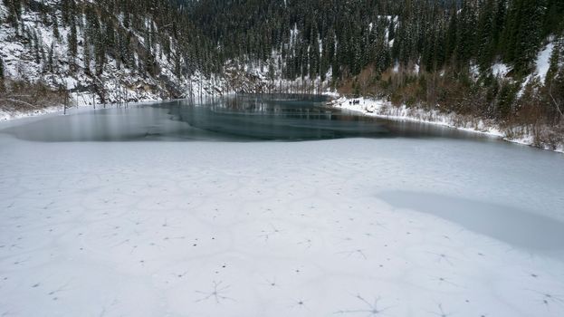 Unusual ice patterns on the mountain lake of Kaindy. Aerial view from a drone of a freezing lake, white ice with round streaks, dark water with a smooth surface. Mountains and coniferous winter forest