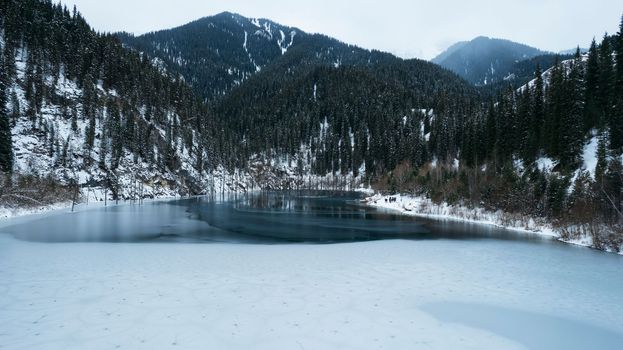 Unusual ice patterns on the mountain lake of Kaindy. Aerial view from a drone of a freezing lake, white ice with round streaks, dark water with a smooth surface. Mountains and coniferous winter forest