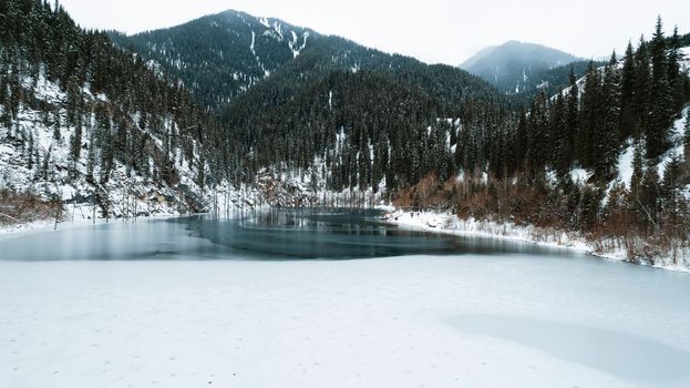 Unusual ice patterns on the mountain lake of Kaindy. Aerial view from a drone of a freezing lake, white ice with round streaks, dark water with a smooth surface. Mountains and coniferous winter forest