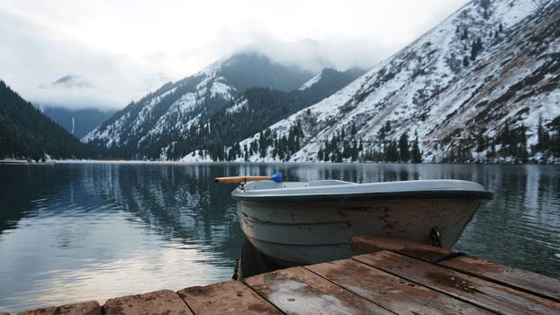 A mountain lake in the forest with mirrored water. Wooden pier with white boat. The water reflects the landscape of a cloudy sky, snowy mountains and peaks, coniferous firs. Kolsai Lake, Kazakhstan