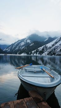 A mountain lake in the forest with mirrored water. Wooden pier with white boats. The water reflects the landscape of a cloudy sky, snowy mountains and peaks, coniferous firs. Kolsai Lake, Kazakhstan