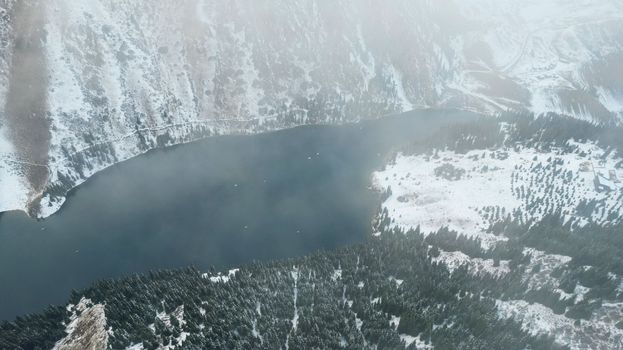 Flying in the clouds over the mountain lake Kolsai. Top view of snowy mountains and forest. Dark lake water. White clouds float by. Steep cliffs in the gorge. Nature of Kazakhstan, Almaty