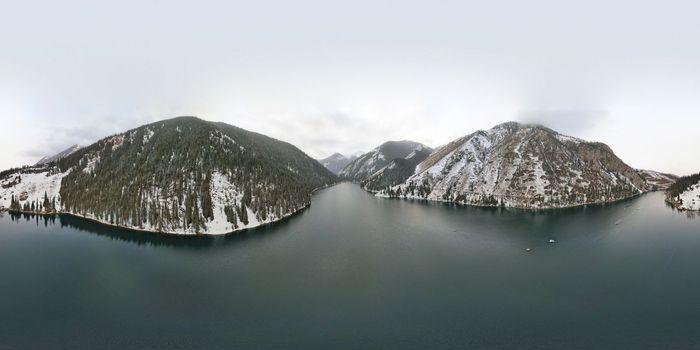 Kolsai mountain lake in the winter forest. Drone view of clouds, coniferous trees, mirrored smooth water, hills and mountains in the snow. Yellow sunset. Boats float in places. Kazakhstan, Almaty