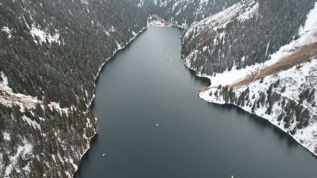 Kolsai mountain lake in the winter forest. Drone view of clouds, coniferous trees, mirrored smooth water, hills and mountains in the snow. Yellow sunset. Boats float in places. Kazakhstan, Almaty