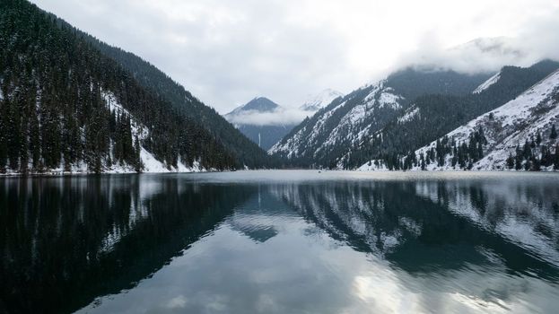 Kolsai mountain lake in the winter forest. Drone view of clouds, coniferous trees, mirrored smooth water, hills and mountains in the snow. Yellow sunset. Boats float in places. Kazakhstan, Almaty