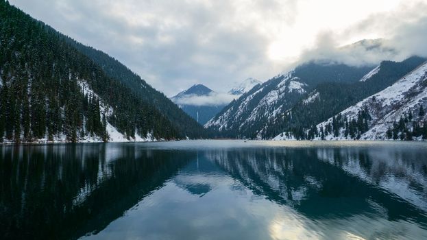 Kolsai mountain lake in the winter forest. Drone view of clouds, coniferous trees, mirrored smooth water, hills and mountains in the snow. Yellow sunset. Boats float in places. Kazakhstan, Almaty