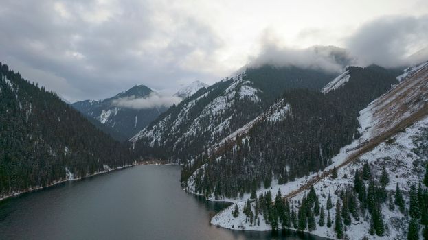 Kolsai mountain lake in the winter forest. Drone view of clouds, coniferous trees, mirrored smooth water, hills and mountains in the snow. Yellow sunset. Boats float in places. Kazakhstan, Almaty
