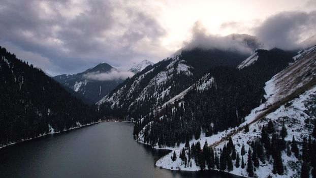 Kolsai mountain lake in the winter forest. Drone view of clouds, coniferous trees, mirrored smooth water, hills and mountains in the snow. Yellow sunset. Boats float in places. Kazakhstan, Almaty