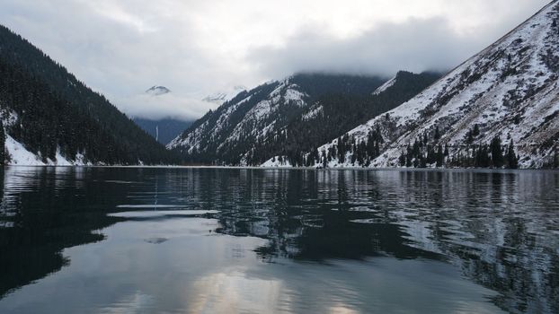 Kolsai mountain lake in the winter forest. Drone view of clouds, coniferous trees, mirrored smooth water, hills and mountains in the snow. Yellow sunset. Boats float in places. Kazakhstan, Almaty