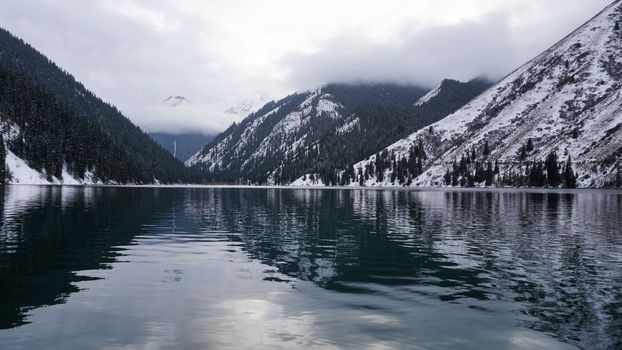 Kolsai mountain lake in the winter forest. Drone view of clouds, coniferous trees, mirrored smooth water, hills and mountains in the snow. Yellow sunset. Boats float in places. Kazakhstan, Almaty