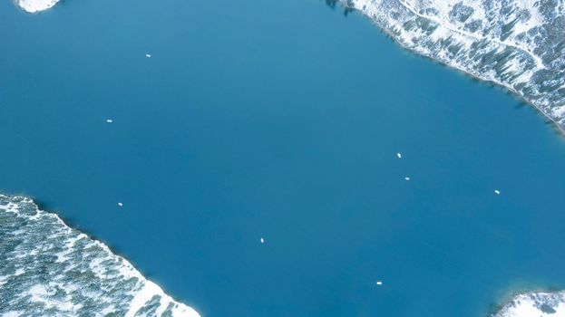 Kolsai mountain lake in the winter forest. Drone view of clouds, coniferous trees, mirrored smooth water, hills and mountains in the snow. Yellow sunset. Boats float in places. Top. Kazakhstan, Almaty