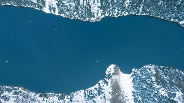 Kolsai mountain lake in the winter forest. Drone view of clouds, coniferous trees, mirrored smooth water, hills and mountains in the snow. Yellow sunset. Boats float in places. Top. Kazakhstan, Almaty