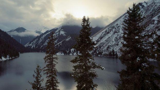 Kolsai mountain lake in the winter forest. Drone view of coniferous trees, mirror-smooth water like a mirror, hills, mountains in snow and clouds. Yellow sunset. Boats float in places. Kazakhstan