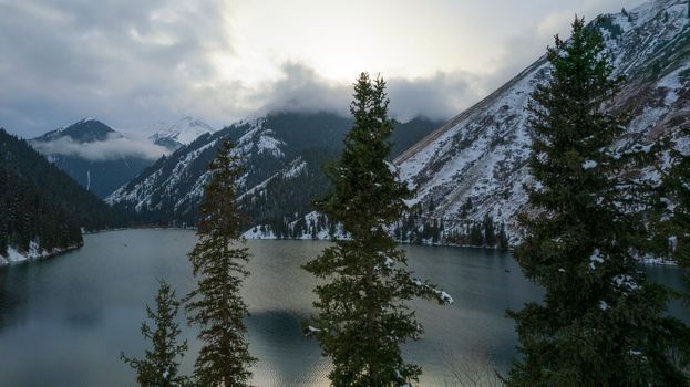 Kolsai mountain lake in the winter forest. Drone view of coniferous trees, mirror-smooth water like a mirror, hills, mountains in snow and clouds. Yellow sunset. Boats float in places. Kazakhstan