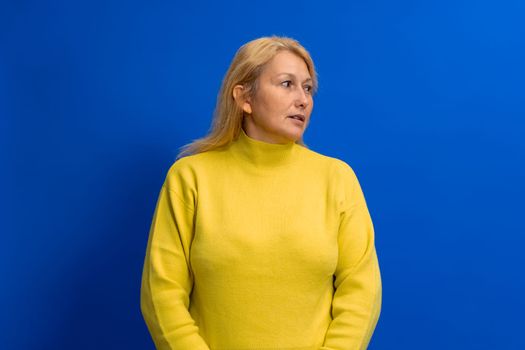 Beautiful woman in her 40s wearing yellow sweater standing on blue background looking to side with serious face