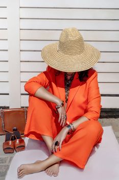 Stylish woman in an orange suit with a hat sits on a rug on a white striped background. On the hands are jewelry rings and bracelets, sandals and a bag stand side by side