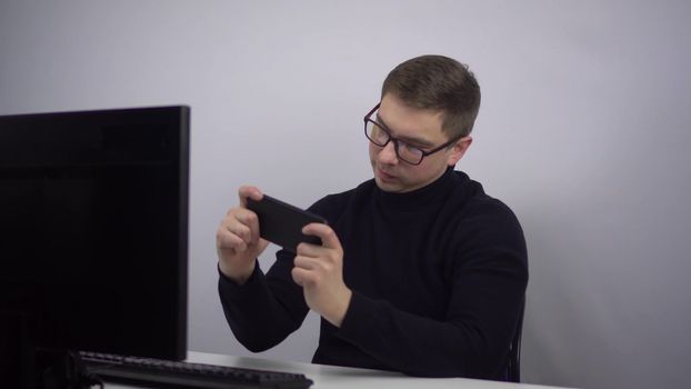 The young man is enthusiastically playing his smartphone and emotionally rejoices at the victory. IT specialist in glasses with a phone in his hands at the workplace in the office. 4k